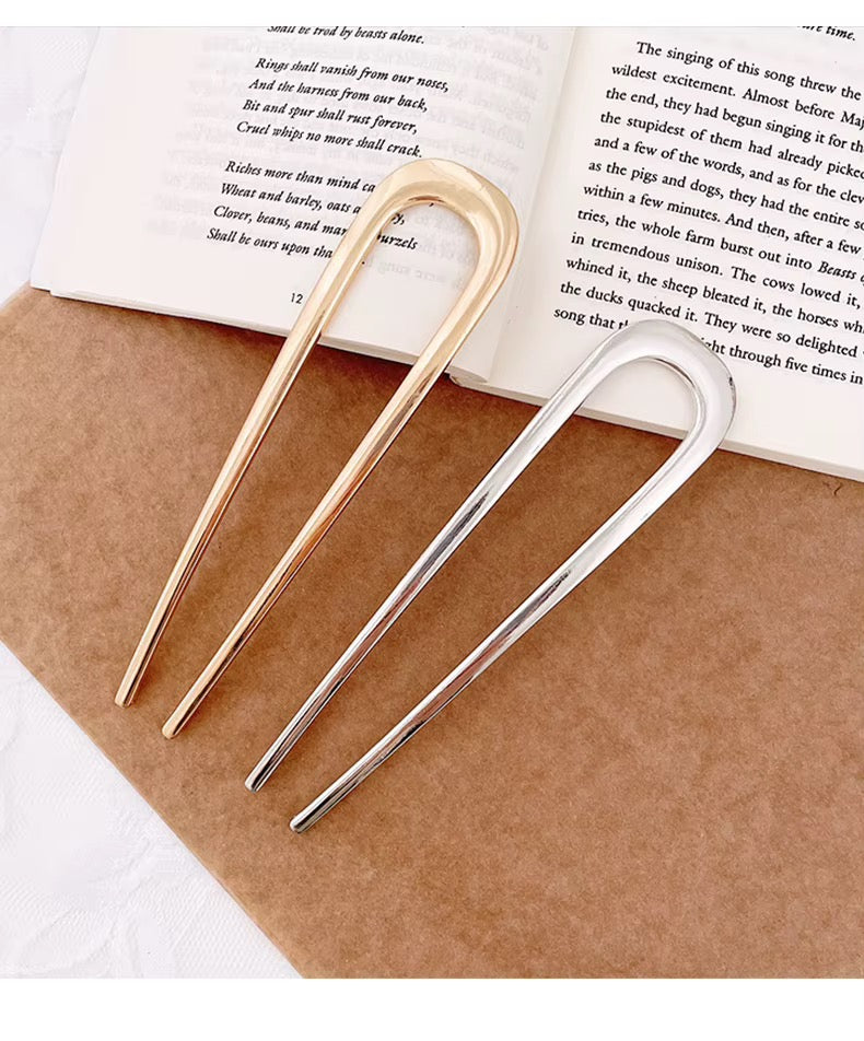 Simply Golden minimalist gold-tone hair forks displayed on a book, showcasing their sleek double-prong construction.