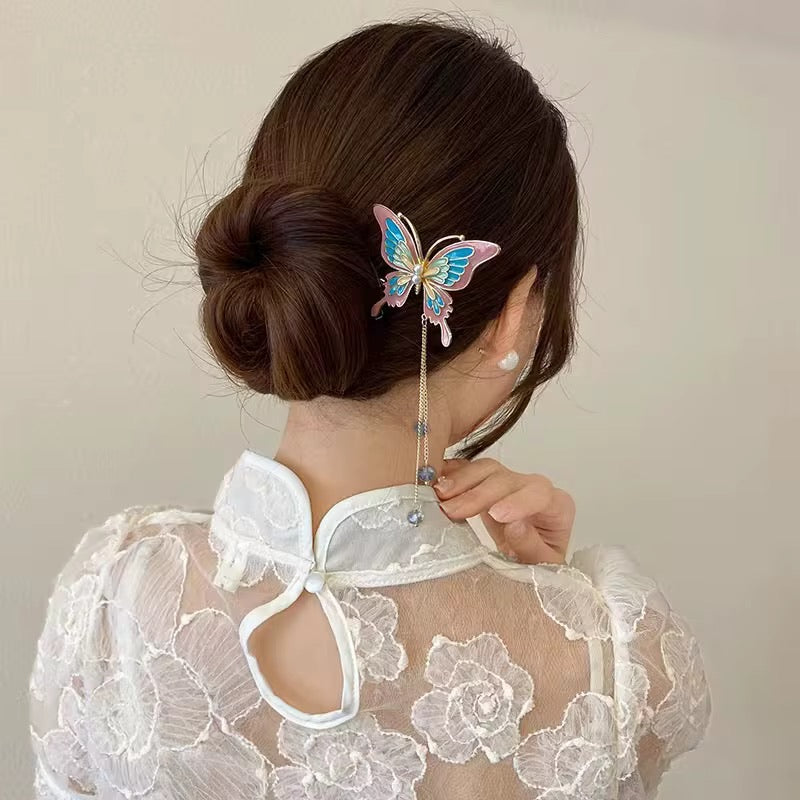 Pastel butterfly hair stick featuring delicate blue and pink wings, a gold-tone stem, and dangling decorative beads, securing an elegant brown hair bun.