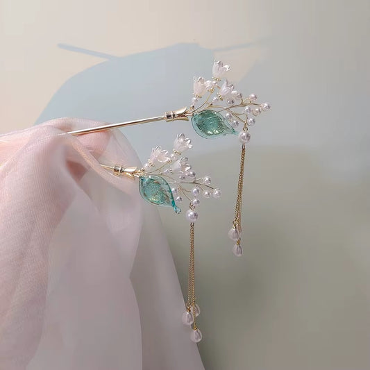 Two matching gold-tone floral hair sticks featuring white bell-shaped blossoms, translucent green leaves, and delicate dangling pearl-like accents.