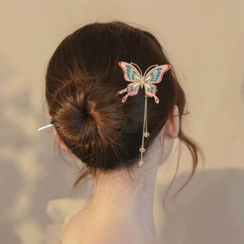 Pastel pink and turquoise butterfly hair stick with gold-tone detailing and dangling crystal-like beads, styled in a brown hair bun with a white lace dress.