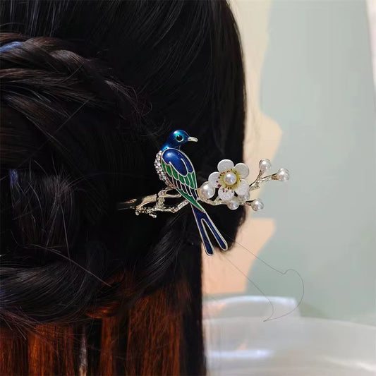 Arayah decorative bird hair stick featuring blue and green enamel detailing, white flowers, and pearl-like accents, styled in a dark twisted updo.