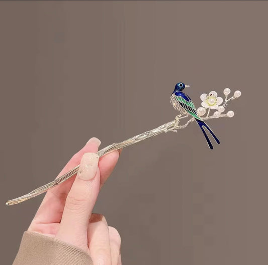 Arayah silver-tone hair stick featuring a blue and green enamel bird perched beside delicate white blossoms and pearl-like accents, held against a neutral background.