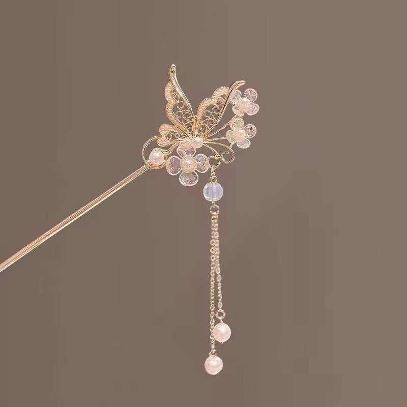 Close-up of a gold-tone butterfly hair stick showcasing intricate crystal-embellished wings, shimmering floral details, and delicate dangling pearl-like accents.