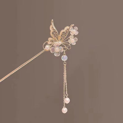 Close-up of a gold-tone butterfly hair stick showcasing intricate crystal-embellished wings, shimmering floral details, and delicate dangling pearl-like accents.