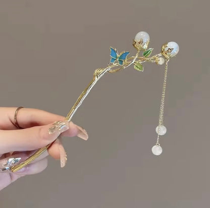 Gold-tone butterfly hair stick featuring a blue butterfly, green leaves, pearl-like embellishments, and dangling decorative beads.