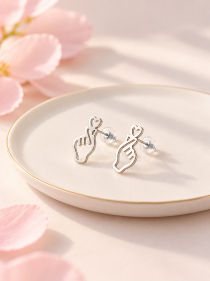 A pair of silver stud earrings shaped like hands holding hearts, displayed on a small round white plate with a gold rim, set against a soft pink background with blurred flower petals.