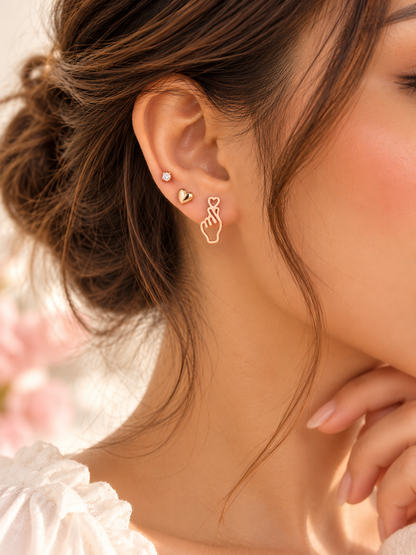 Styling inspiration: an ear wearing a gold-tone finger-heart stud with a small heart stud and a round sparkling stud. Only the Love Gesture Studs pair is included; other earrings are not included.