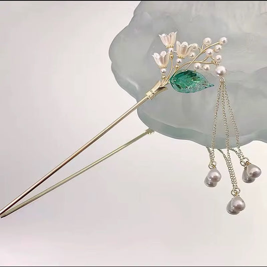 Gold-tone floral hair stick with white blossoms, a translucent green leaf, and dangling pearl-like beads displayed against a pale decorative surface.