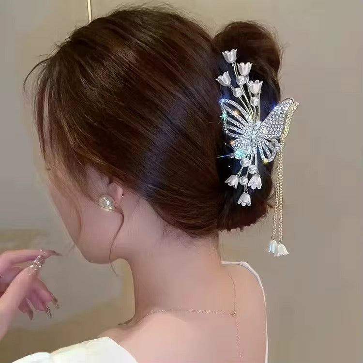 Gold-tone butterfly hair claw clip adorned with sparkling clear rhinestones, ivory tulip-shaped flowers, faux pearl accents, and dangling gold chains with floral charms, securing a voluminous brunette bun in a side-profile styling view.