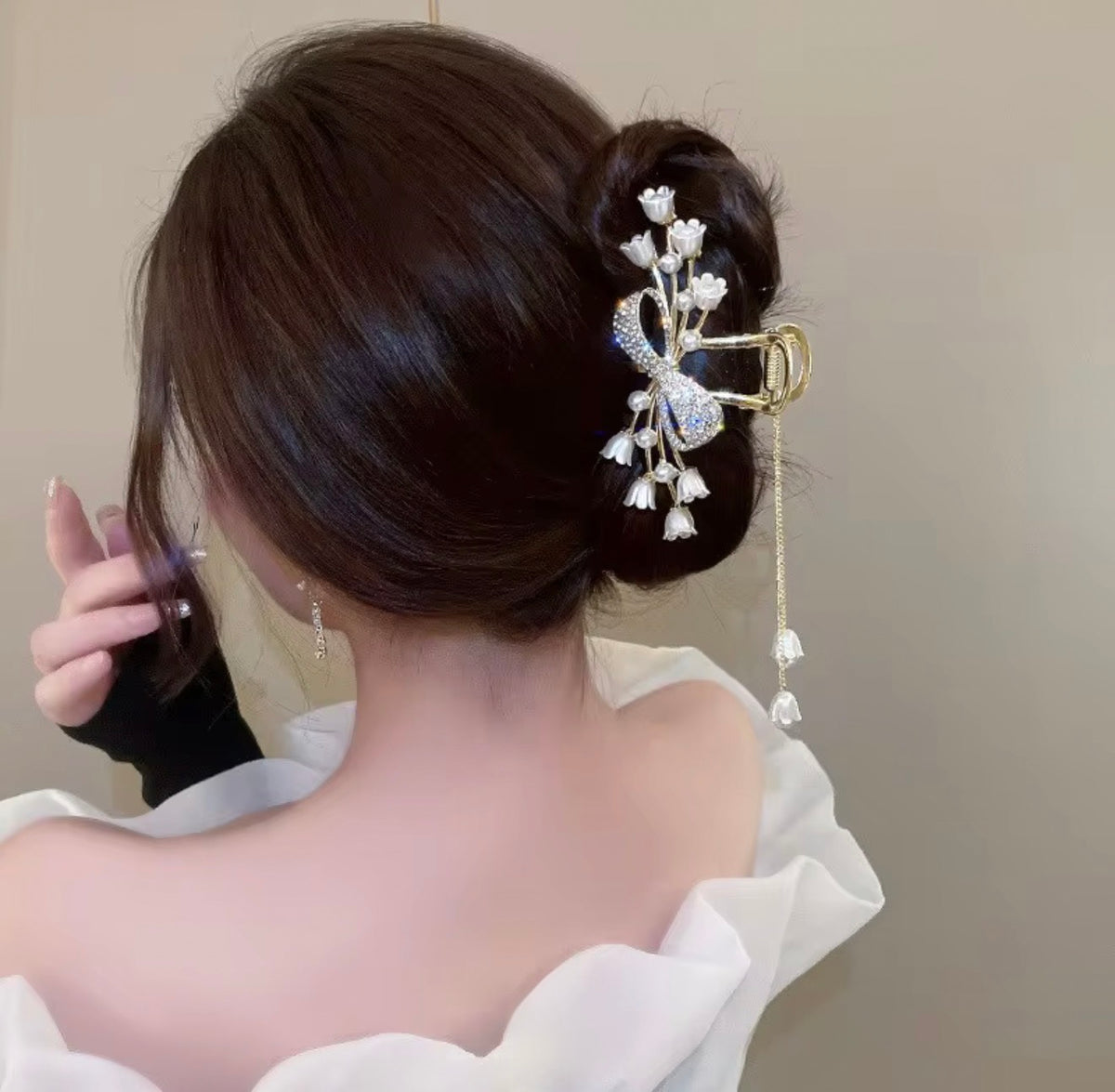Gold-tone bow hair claw clip adorned with sparkling rhinestones, ivory tulip-shaped flowers, and delicate faux pearls, securing a polished brunette bun with dangling gold chains and floral charms.