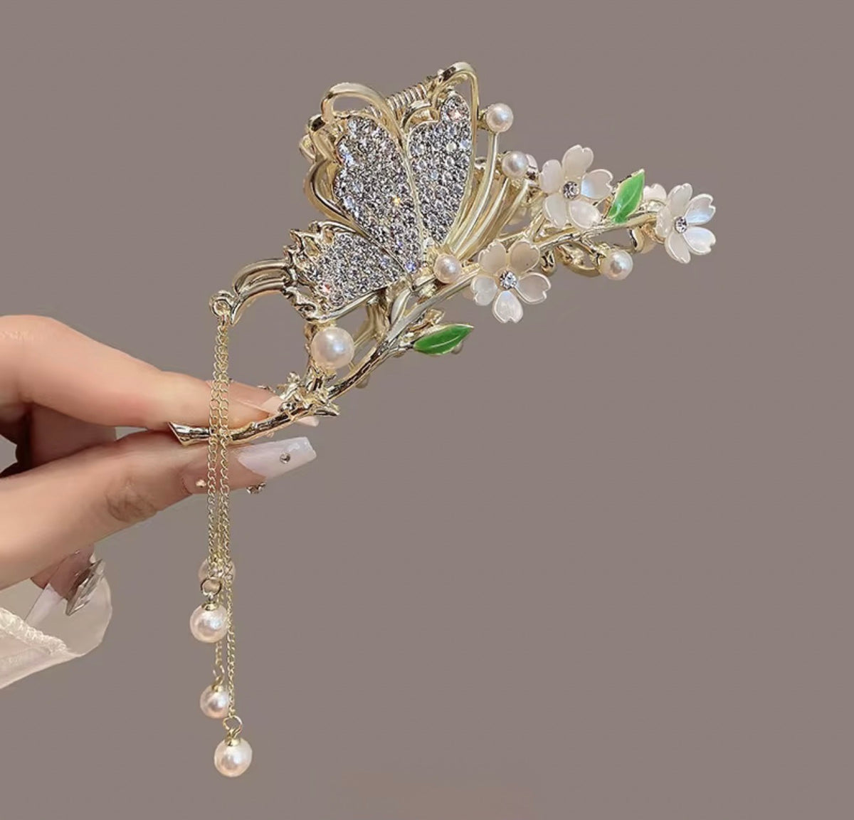 Tulip Blanc Crystal design: gold-tone butterfly hair claw with sparkling wings, white blossoms, green leaf accents, and cascading pearl-style drops, displayed in hand.