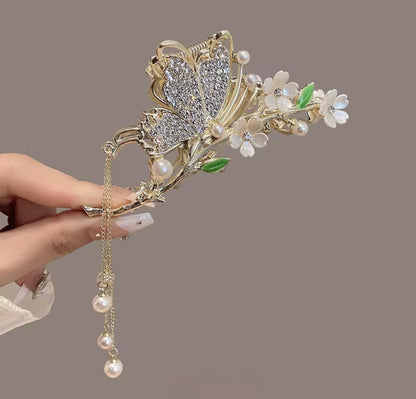 Tulip Blanc Crystal design: gold-tone butterfly hair claw with sparkling wings, white blossoms, green leaf accents, and cascading pearl-style drops, displayed in hand.