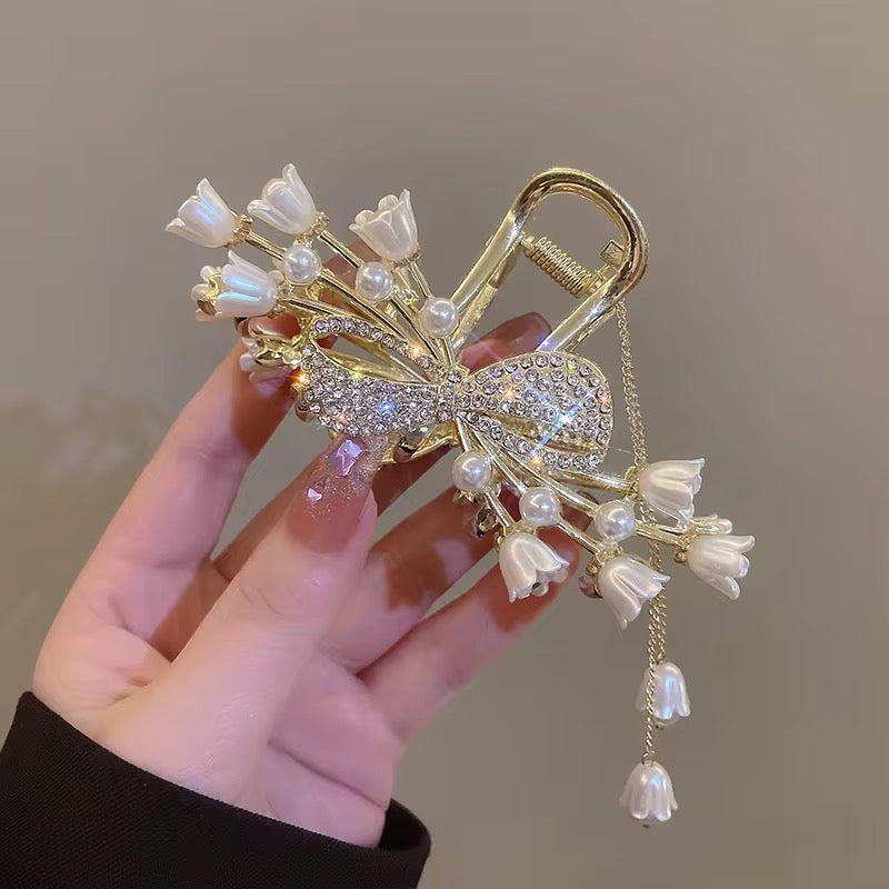 Tulip Blanc Solenne design: gold-tone bow hair claw with sparkling accents, white tulip-shaped flowers, pearl-style details, and dangling floral charms, displayed in hand.