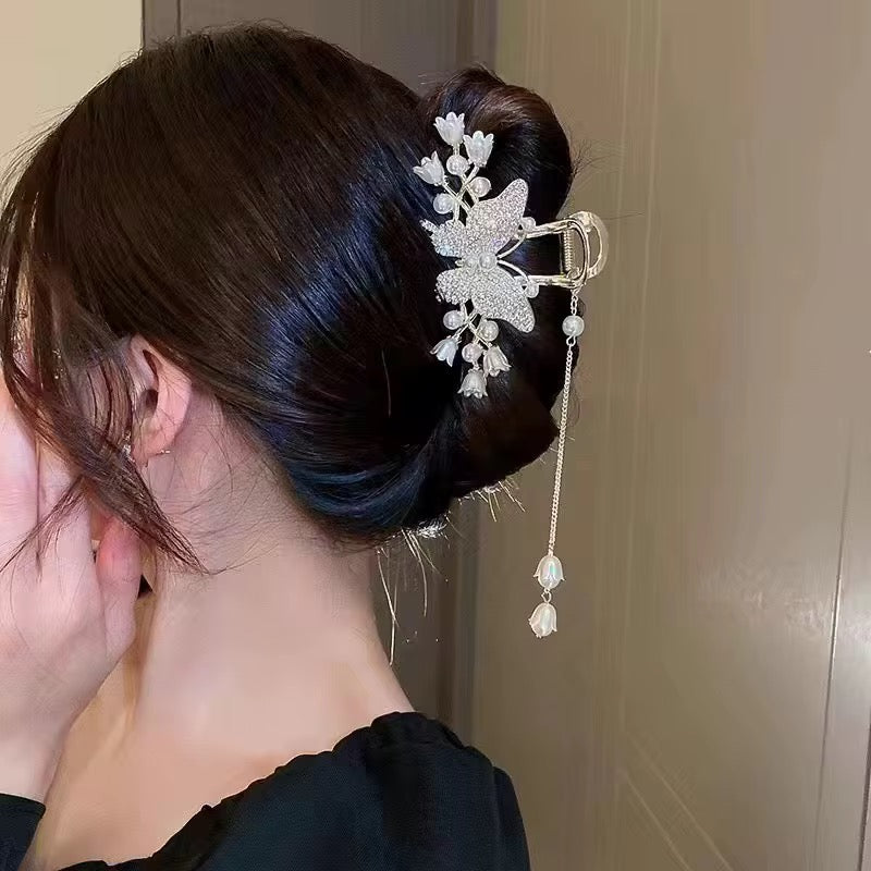 Gold-tone butterfly hair claw clip featuring sparkling rhinestone wings, ivory tulip-shaped flowers, faux pearl accents, and a dangling gold chain with floral charms, securing a sleek brunette low bun in a side-profile styling view.