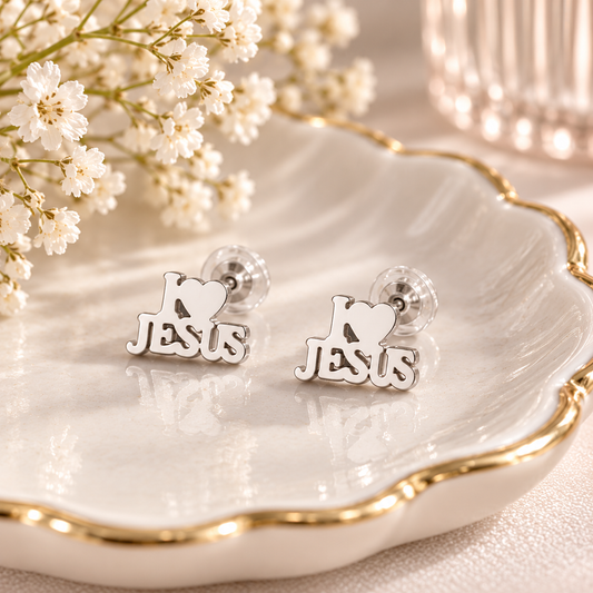 A pair of silver-toned stud earrings shaped like the word Jesus with a heart symbol, displayed on a white scalloped dish with gold trim and small white flowers.