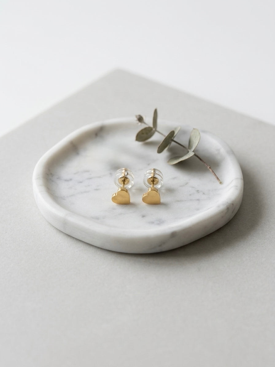 A pair of small gold heart stud earrings displayed on a round white marble tray with a sprig of eucalyptus, set on a light gray surface.