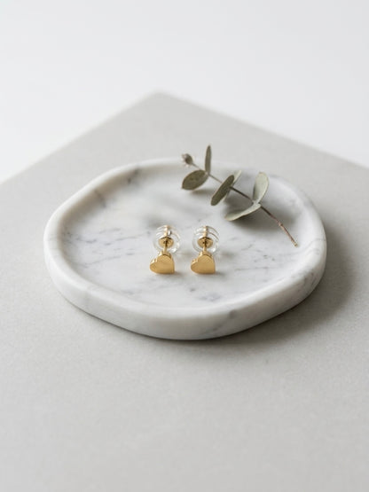 A pair of small gold heart stud earrings displayed on a round white marble tray with a sprig of eucalyptus, set on a light gray surface.