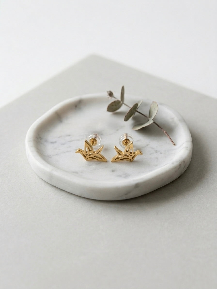 A pair of gold-tone origami crane stud earrings on a round white marble tray beside a sprig of eucalyptus.