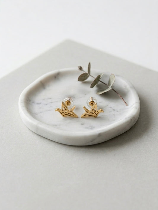 A pair of small gold leaf stud earrings resting on a round white marble tray with a sprig of eucalyptus, set against a neutral gray background.