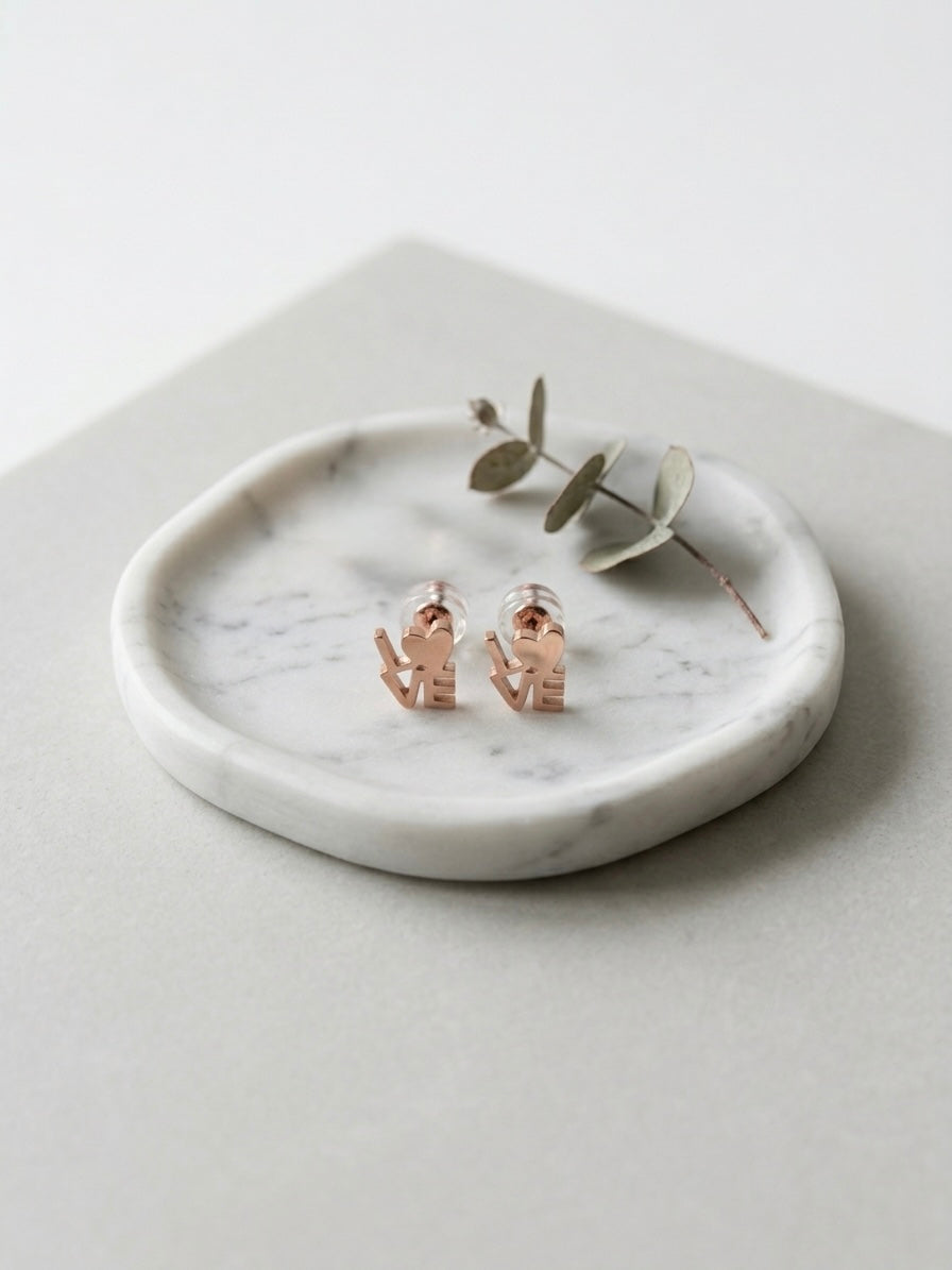 Rose gold-tone LOVE stud earrings with heart-shaped lettering and clear backings, displayed on a white marble dish.