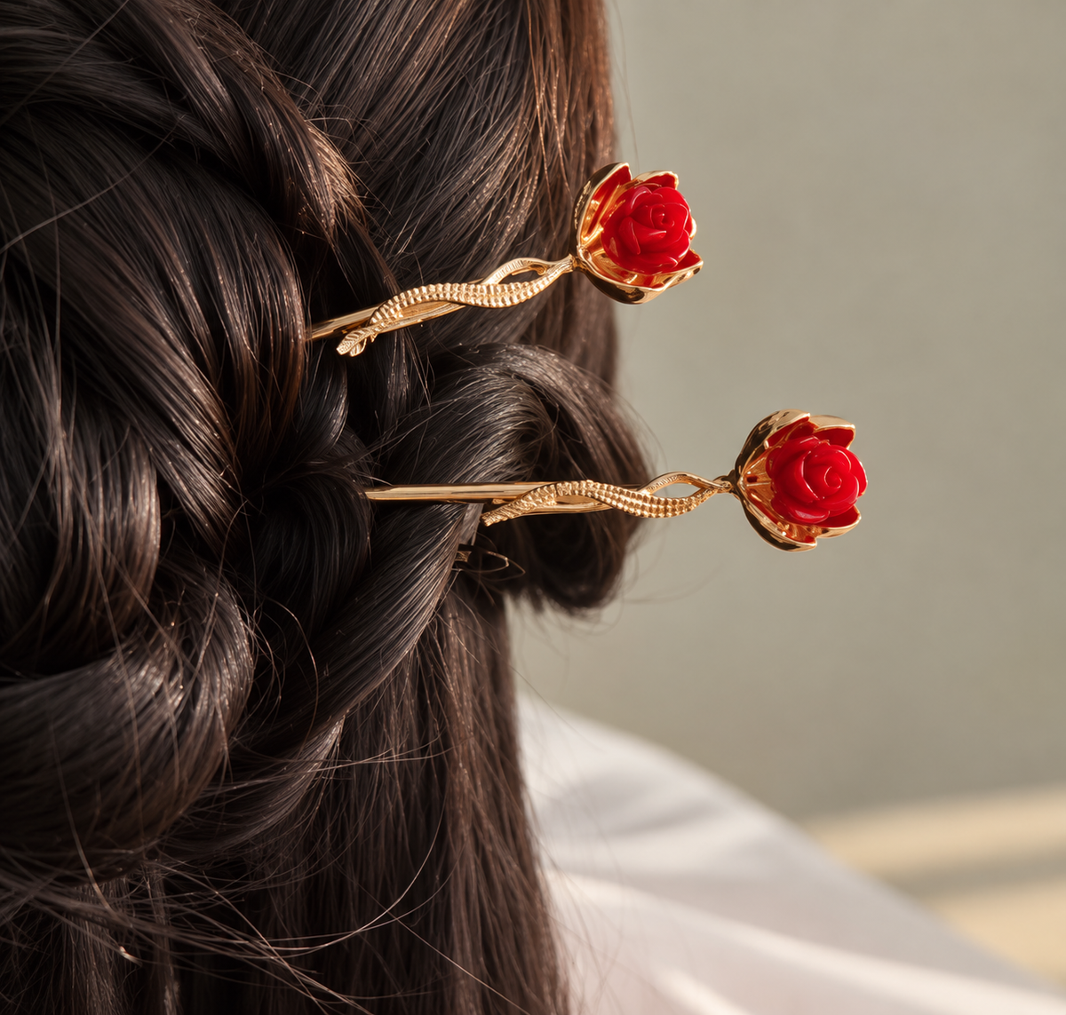 Two gold-tone hair sticks featuring miniature red roses with sculpted petals and delicate twisted stems, styled in a dark brown braided updo.